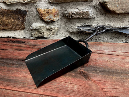 Black metal dustpan on a wooden surface with a stone wall background