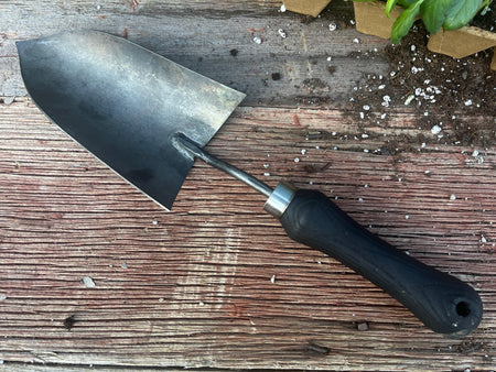 Gardening trowel on a wooden surface with soil and plant pot in the background