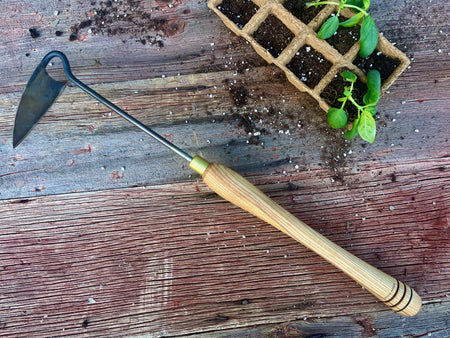 Gardening tool with wooden handle on a wooden surface with soil and plants.